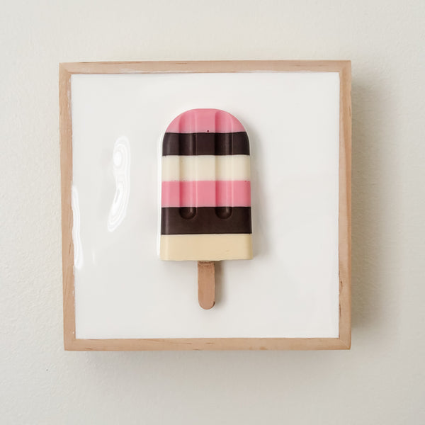 A square framed wall art piece featuring a stylized popsicle with pink, cream, and dark brown horizontal stripes on a white background, mounted on a light beige wall.